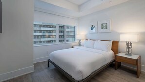Une chambre moderne avec un grand lit, des tables de chevet, une fenêtre donnant sur un immeuble et des œuvres d’art encadrées accrochées au mur.