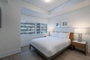 A modern bedroom with a large bed, bedside tables, a window with a view of a building, and framed artwork on the wall.