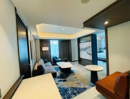 A modern hotel room with a large bed, a sofa, a coffee table, and a small table. There is a view of the city through the window.