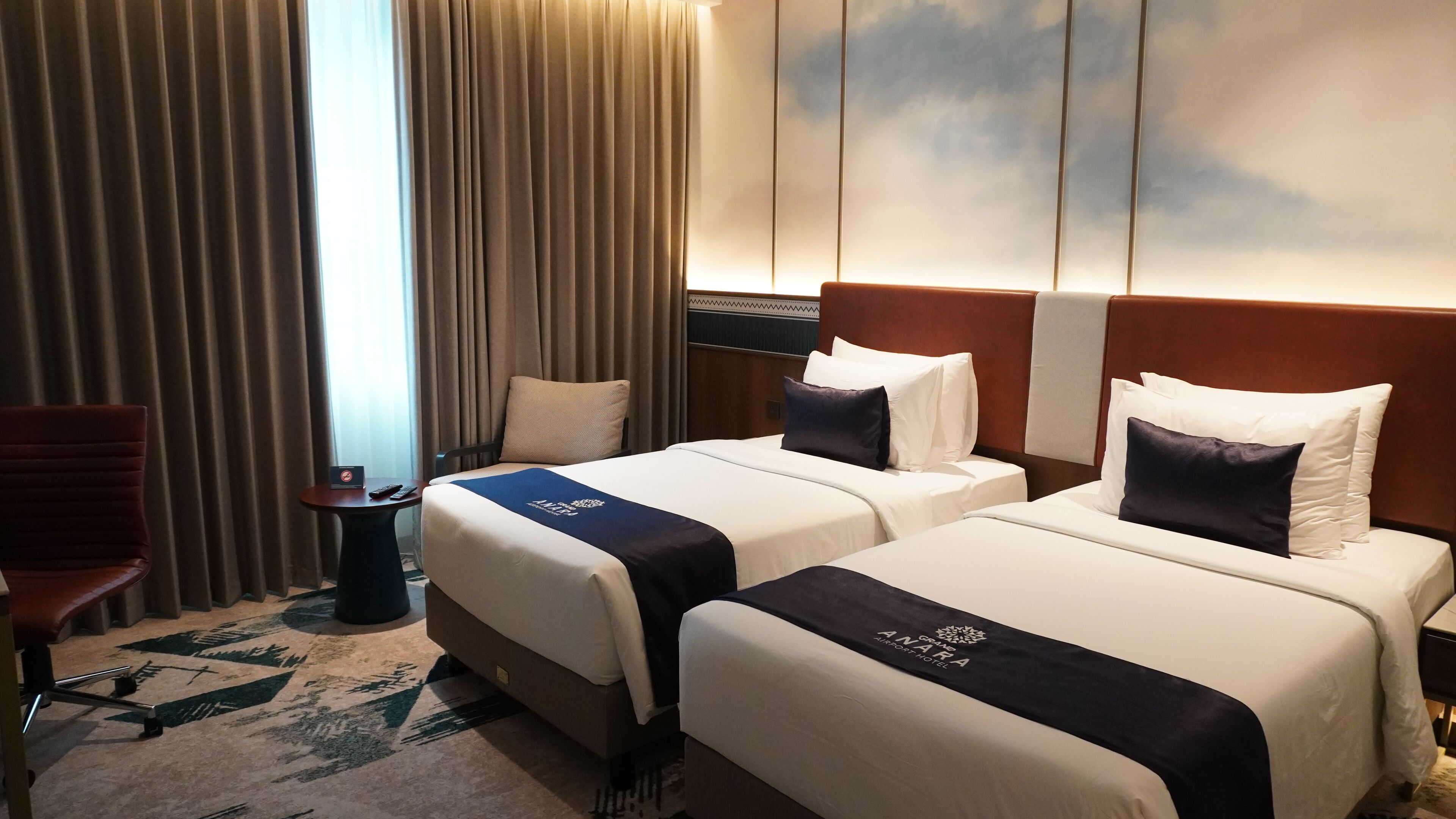 Une chambre d’hôtel avec deux lits, un bureau, une chaise et une grande fenêtre avec des rideaux.