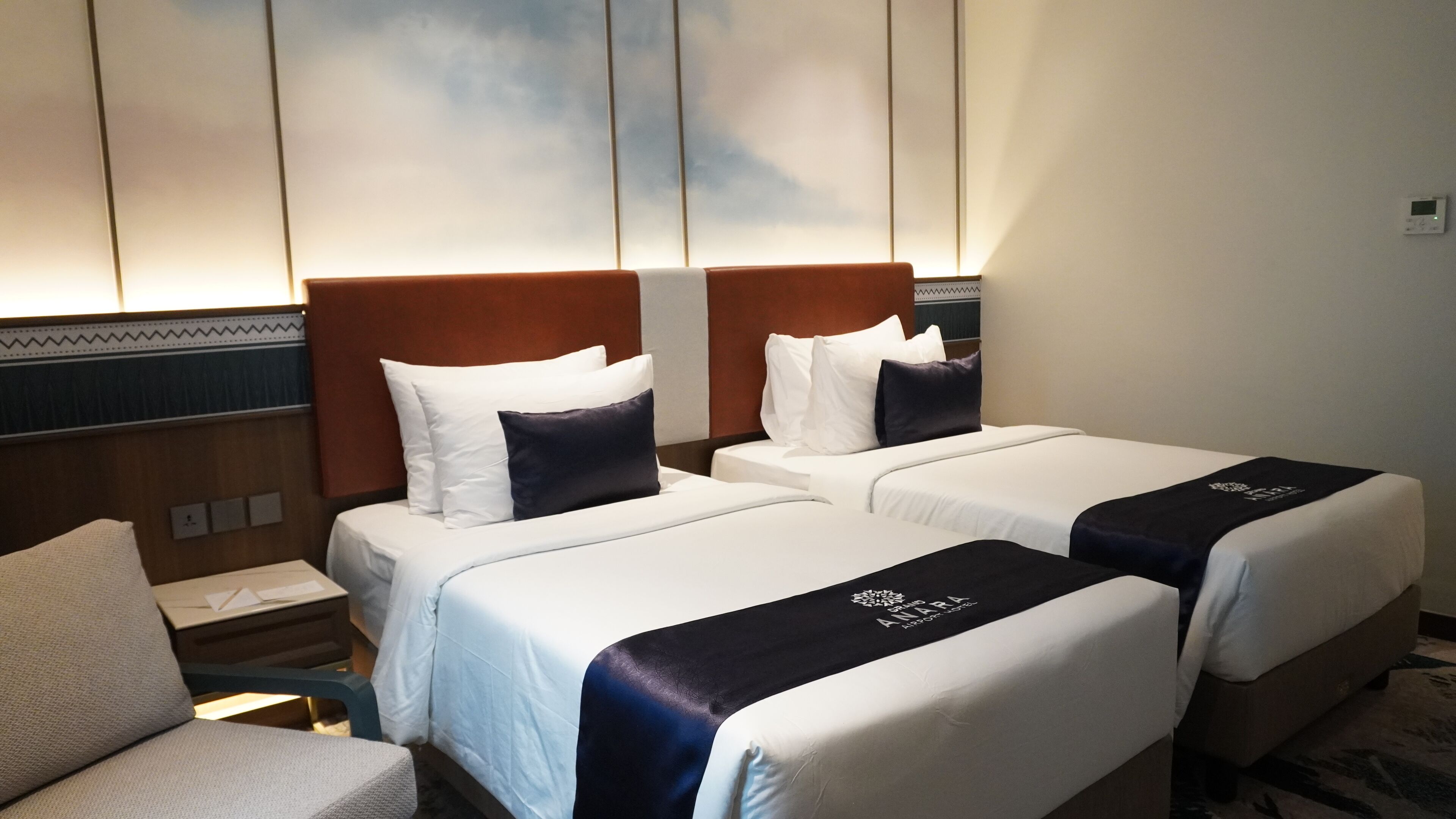 A hotel room with two beds, each with white bedding and dark blue headboards. There is a small desk with a chair and a wall-mounted control panel.