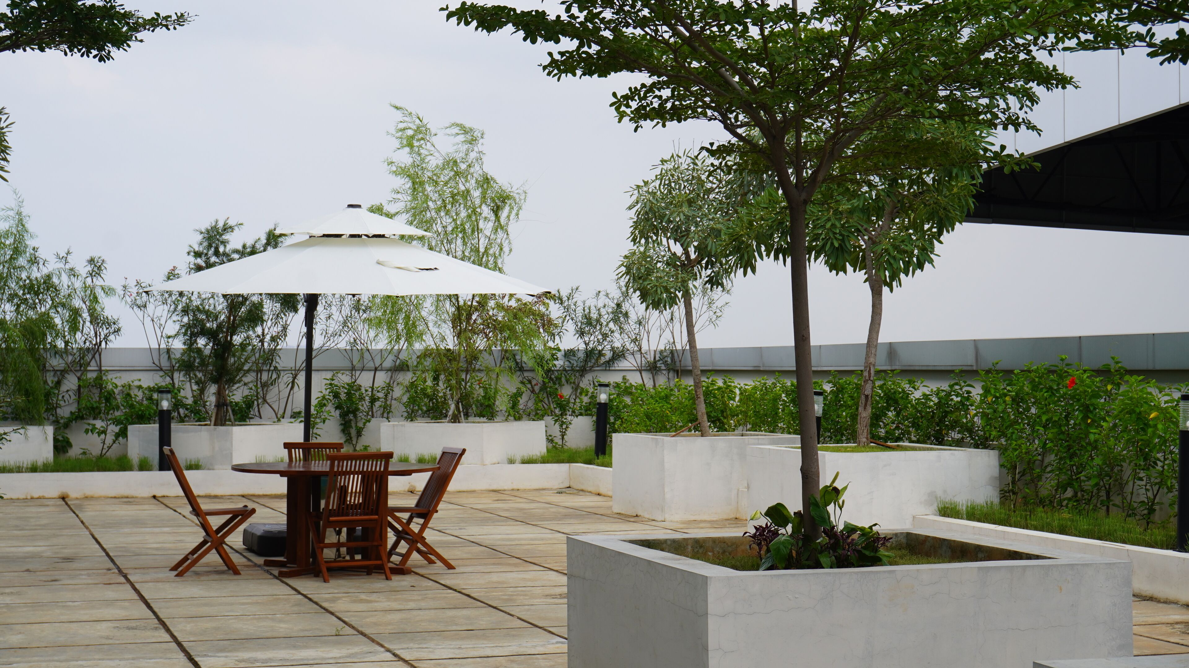 Eine Terrasse im Freien mit Tisch und Stühlen unter einem weißen Sonnenschirm, umgeben von Grünpflanzen und einem modernen Gebäude im Hintergrund.