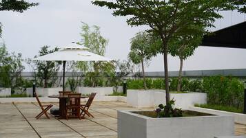 Eine Terrasse im Freien mit Tisch und Stühlen unter einem weißen Sonnenschirm, umgeben von Grünpflanzen und einem modernen Gebäude im Hintergrund.