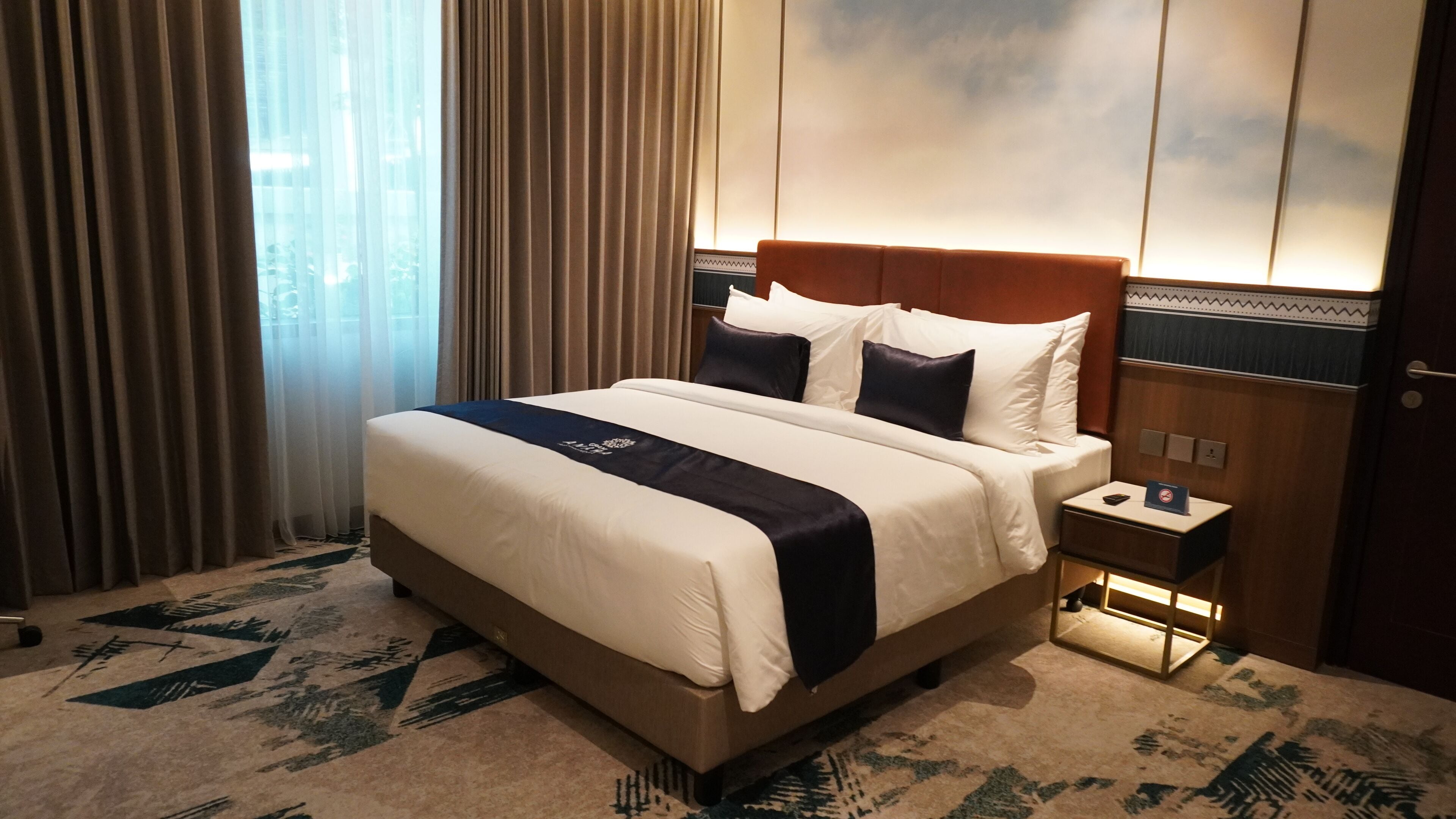 A hotel room with a large bed, a nightstand, a lamp, and a view of the outdoors through the window.