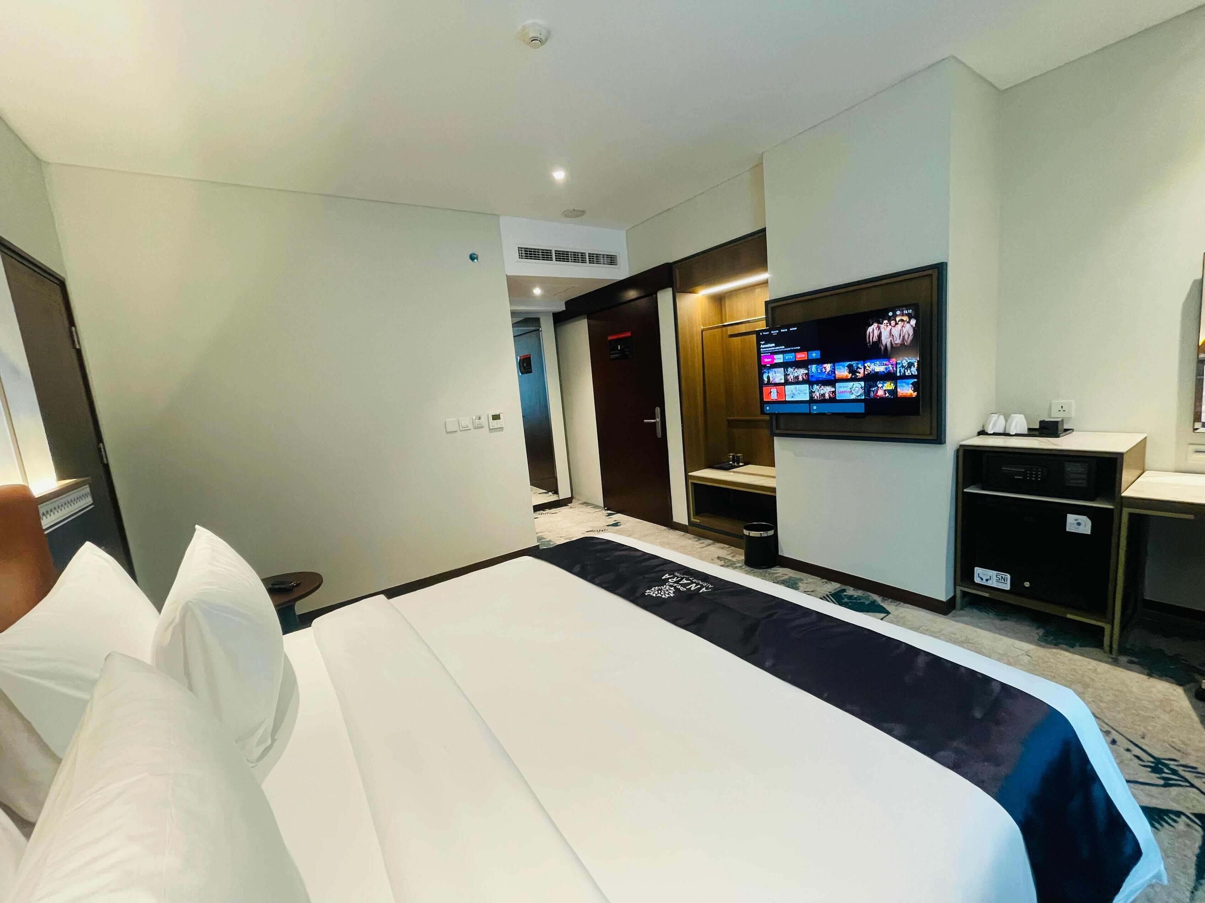 A hotel room with a bed, a TV mounted on the wall, a desk with a computer, and a small trash bin.