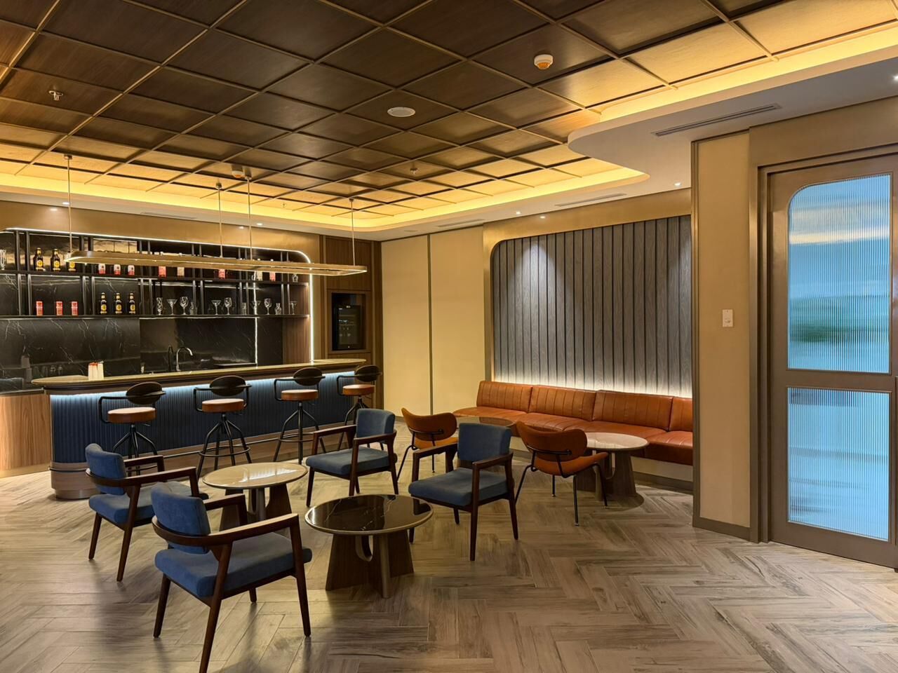 A modern bar area with a wooden floor, blue and brown seating, and a bar counter with bottles and glasses.