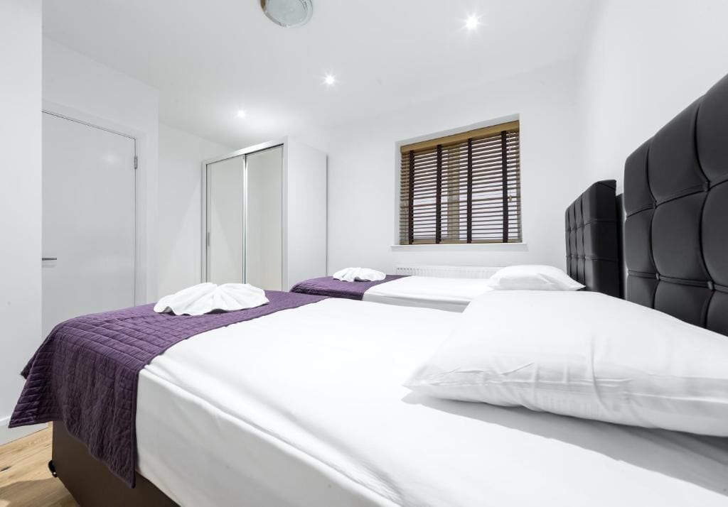 A modern hotel room with two beds, a large window with blinds, and a dark headboard.