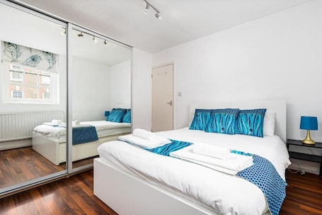 A modern hotel room with a large bed, a mirror, a nightstand, and a window with a view of buildings.