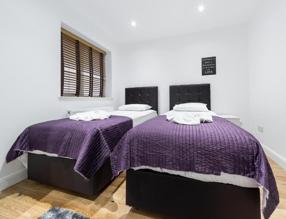 Een slaapkamer met twee bedden, elk met een zwart hoofdeinde en paars beddengoed. Er is een raam met zonwering, een witte muur en een bord met een citaat.