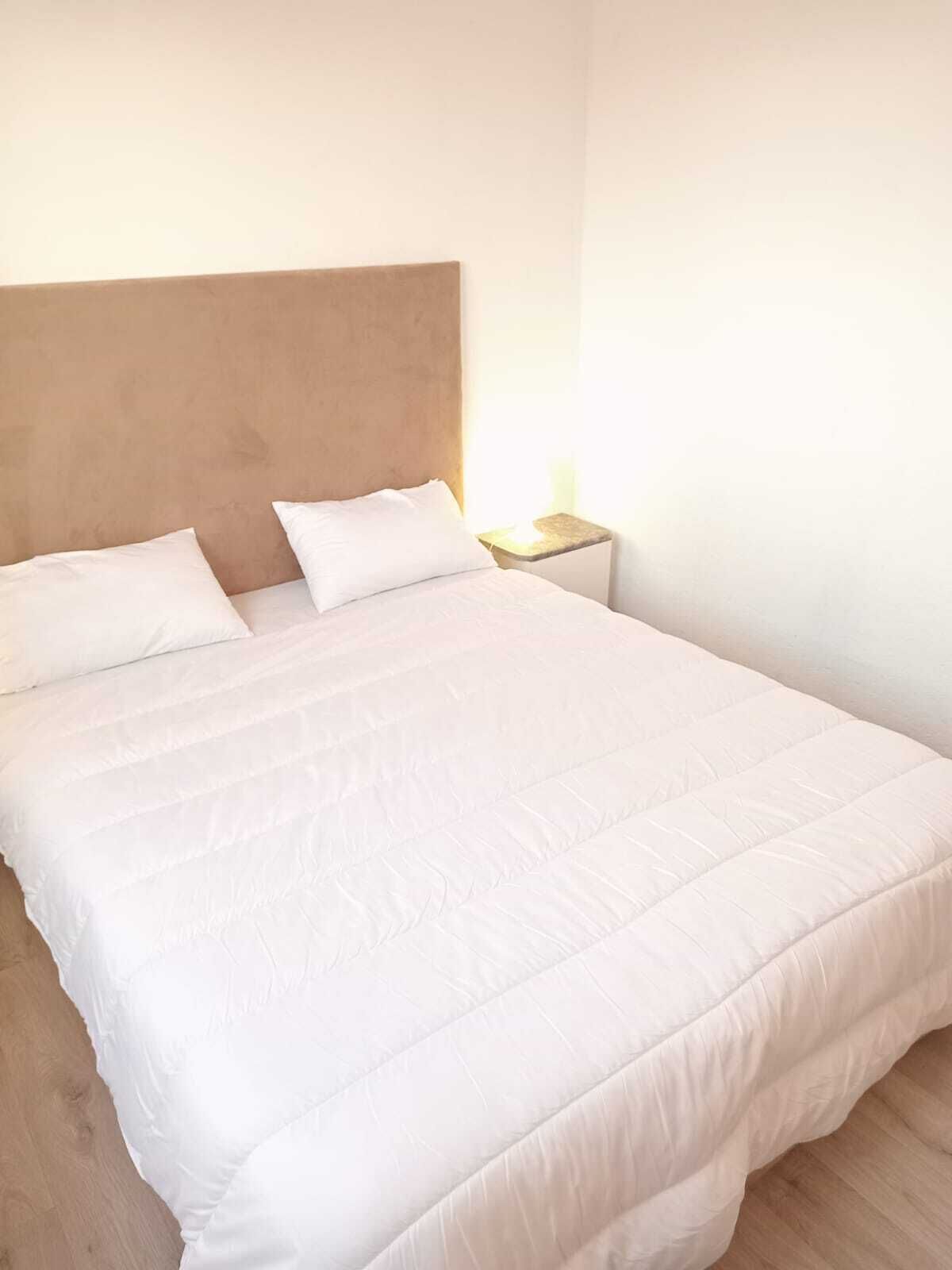 Een net opgemaakt bed met witte lakens in een kamer met een houten hoofdbord en een nachtkastje met een lamp.