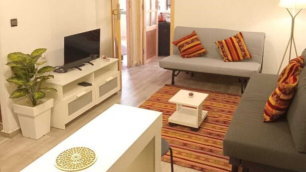 Un salon moderne avec un canapé gris, un meuble TV blanc et une petite table à manger avec deux chaises. Une plante en pot se trouve à gauche, et une lampe à droite.