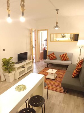 Ein modernes Wohnzimmer mit grauem Sofa, einem weißen TV-Schrank und einem kleinen Esstisch mit zwei Stühlen. Eine Kühlpflanze steht links, und rechts befindet sich eine Lampe.
