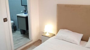 Une chambre d’hôtel comprenant un lit, une table de chevet avec une lampe, une petite salle de bain avec un lavabo et un miroir, et une porte donnant sur une autre pièce.