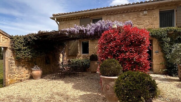 Un bâtiment en pierre avec une cour recouverte de gravier, des plantes en pot et une pergola couverte de fleurs.