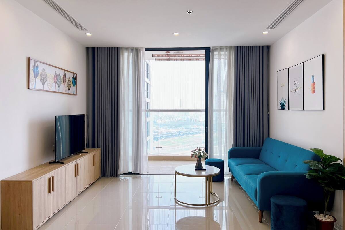 A modern living room with a blue sofa, a TV on a wooden cabinet, a small round table, and a large window with curtains.