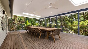 A covered outdoor dining area with a wooden table and chairs, surrounded by lush greenery.