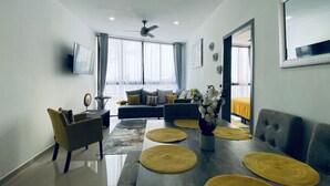 Ein modernes Wohnzimmer mit einer Couch, einem Esstisch mit Platzdeckchen, einem an der Wand montierten Fernseher und einem Deckenventilator.