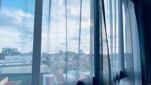 Blick durch ein Fenster mit transparenten Vorhängen auf eine Stadtlandschaft mit Gebäuden und blauem Himmel.