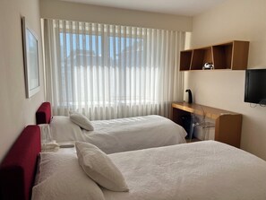 Una camera d'albergo con due letti, una scrivania, una televisione e una finestra con tende leggere.