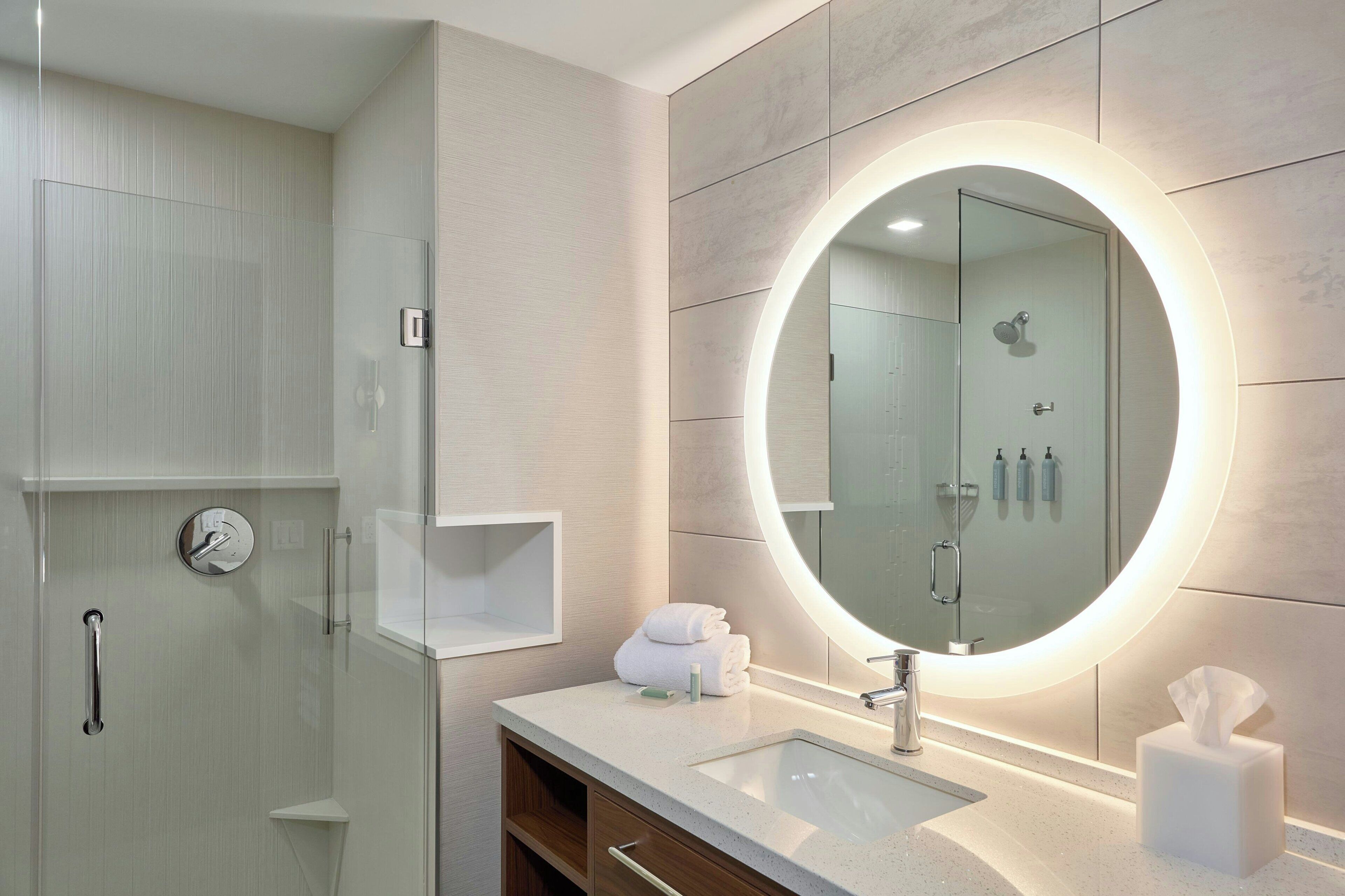 A modern bathroom with a glass shower enclosure, a vanity with a sink, and a large mirror with a circular light.