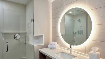 A modern bathroom with a glass shower enclosure, a vanity with a sink, and a large mirror with a circular light.
