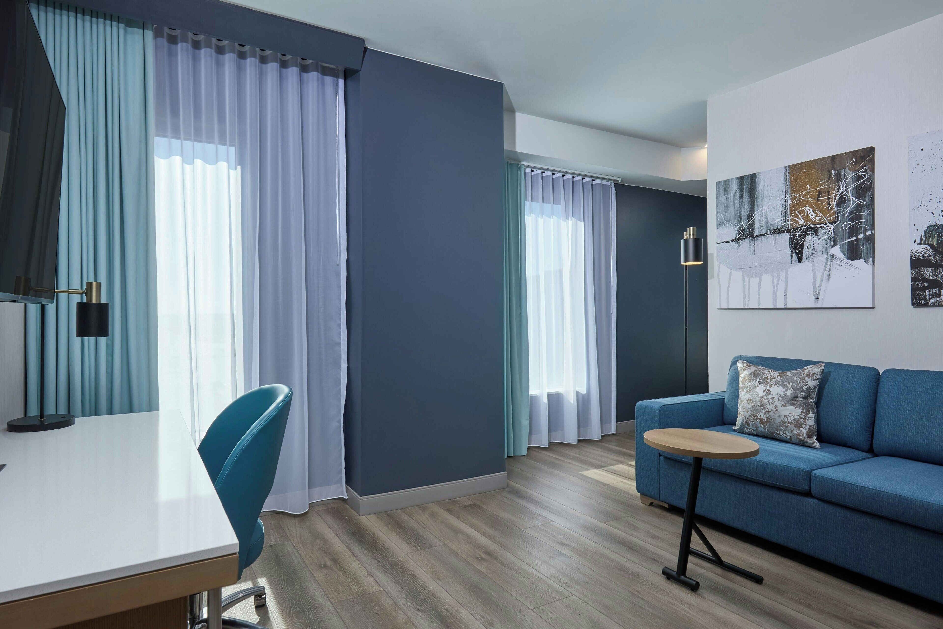 Un salon moderne avec un canapé bleu, une table basse ronde et un fauteuil bleu. On y trouve un bureau blanc avec une lampe, ainsi qu’une grande fenêtre ornée de rideaux légers.