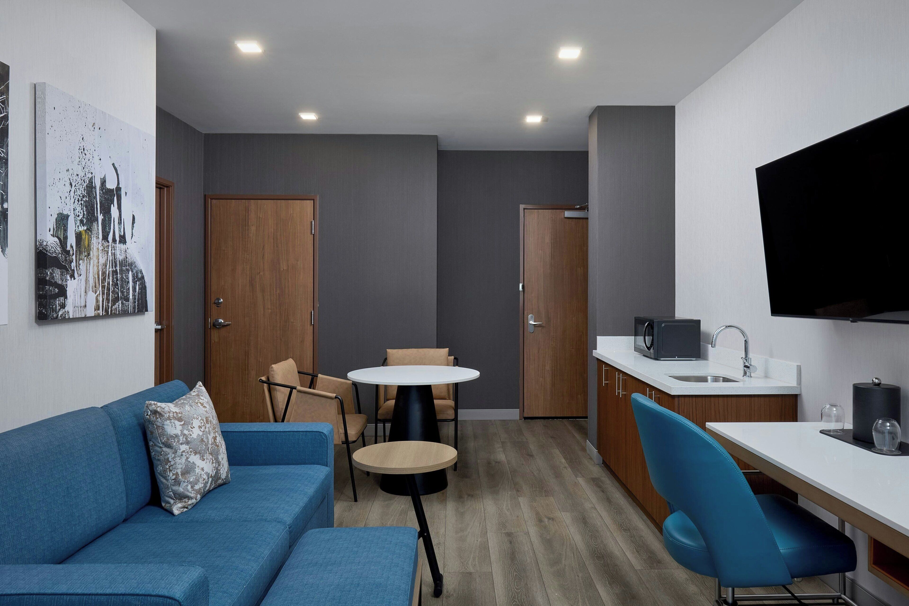 A compact, modern room with a blue sofa, a round table, a blue chair, a TV, and a kitchenette.