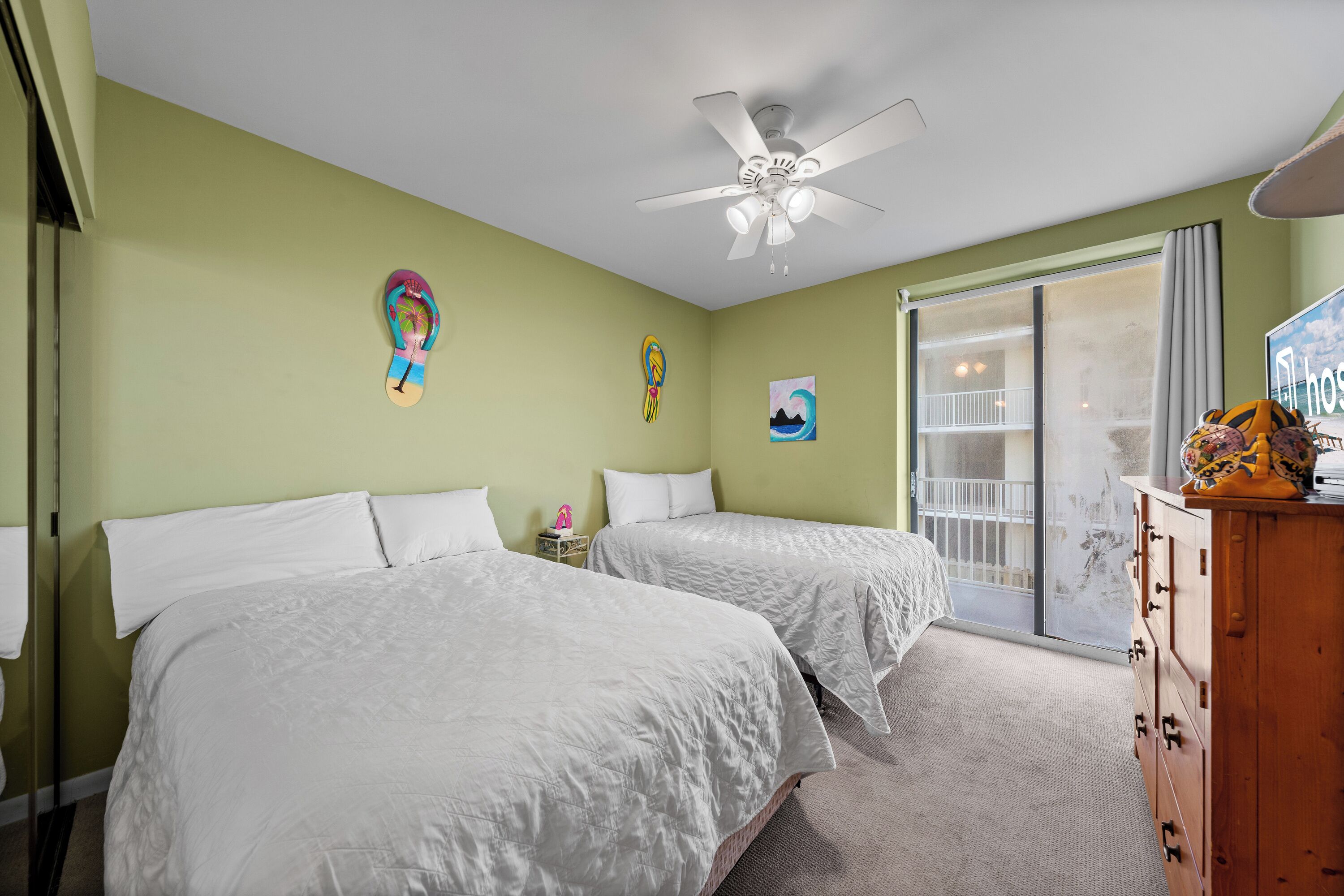A hotel room with two beds, a ceiling fan, a dresser, and a view of the outside balcony.
