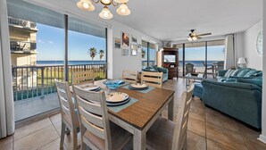 Un vaste espace de vie comprenant une table à manger, des chaises et un canapé. Un balcon offre une vue sur l’océan et les palmiers.
