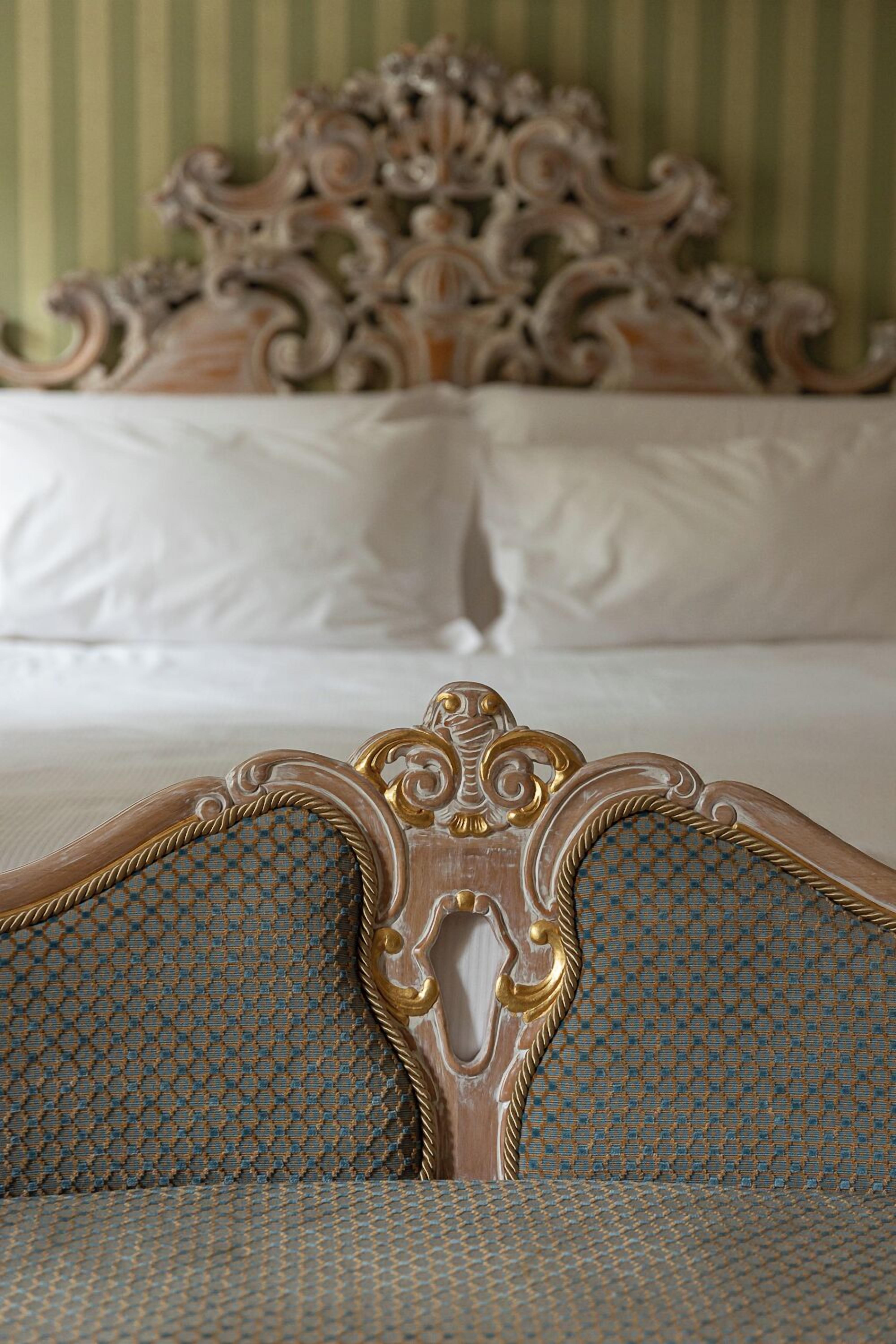 A bed with ornate headboard, a patterned upholstered chair, and a striped wall.