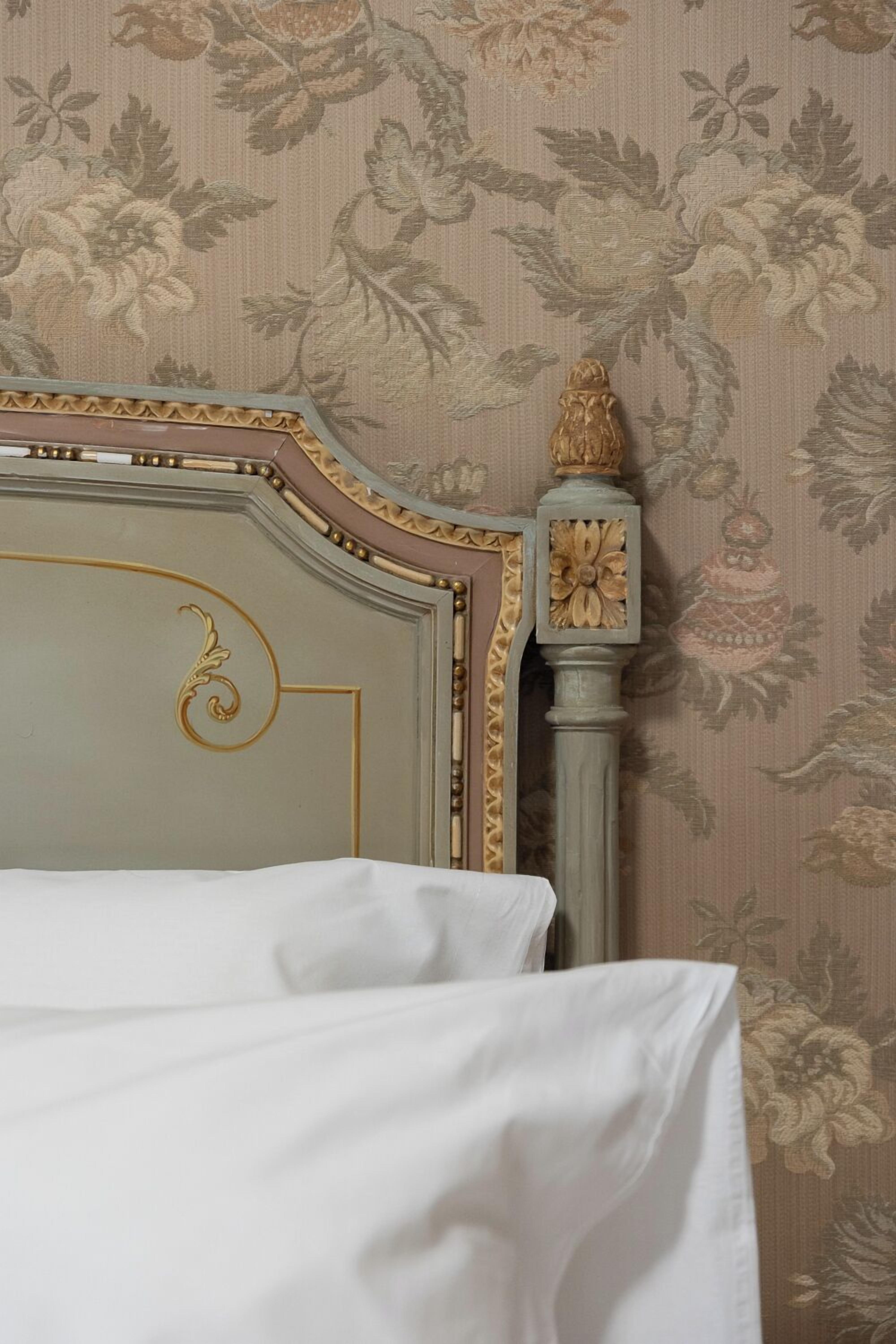 Ein Bett mit weißer Bettdecke, einem verzierten Kopfteil mit Goldzierde und floralem Wandpapier.