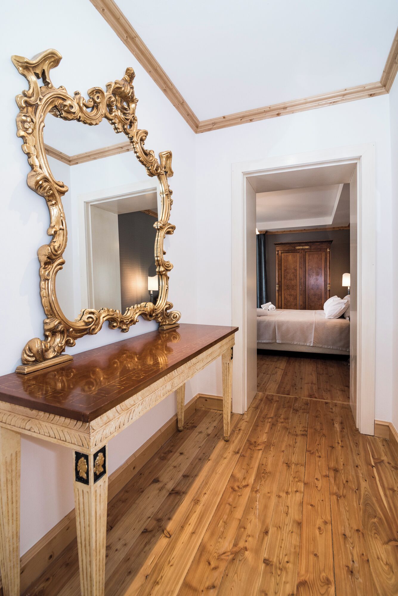 Une pièce avec un plancher en bois, un grand miroir décoratif posé sur une table en bois, et un lit avec une tête de lit sombre à l’arrière-plan.