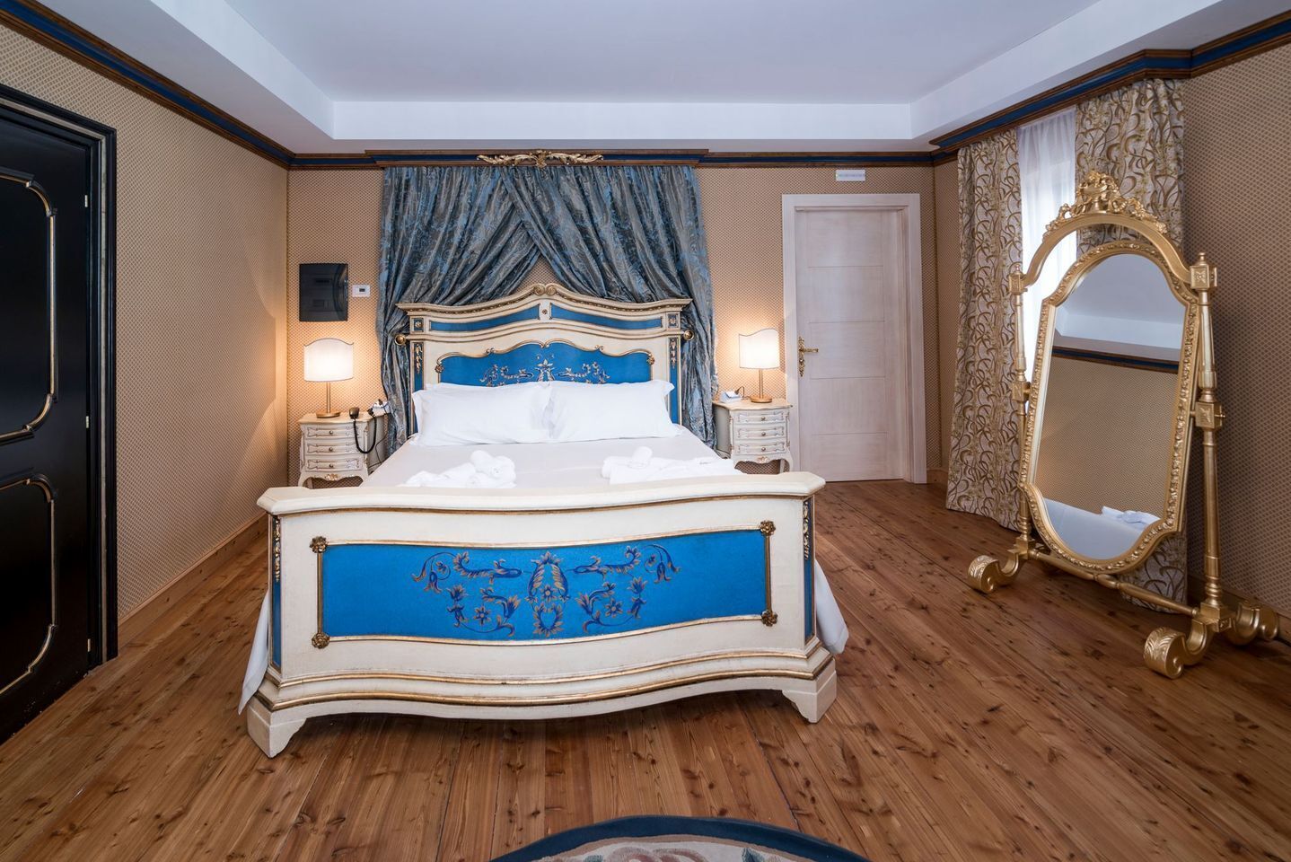 Een groot bed met versierde hoofdbord en voetenbord, twee nachtkastjes, een grote spiegel en een deur in een kamer met houten vloer en behang met patroon.