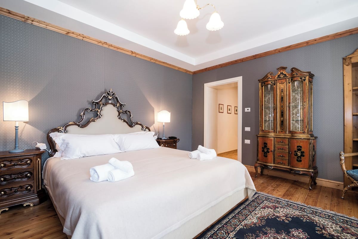 Habitación de hotel con una cama grande, cabecera de madera, dos lámparas de noche, cómoda de madera, un espejo grande y una alfombra estampada.