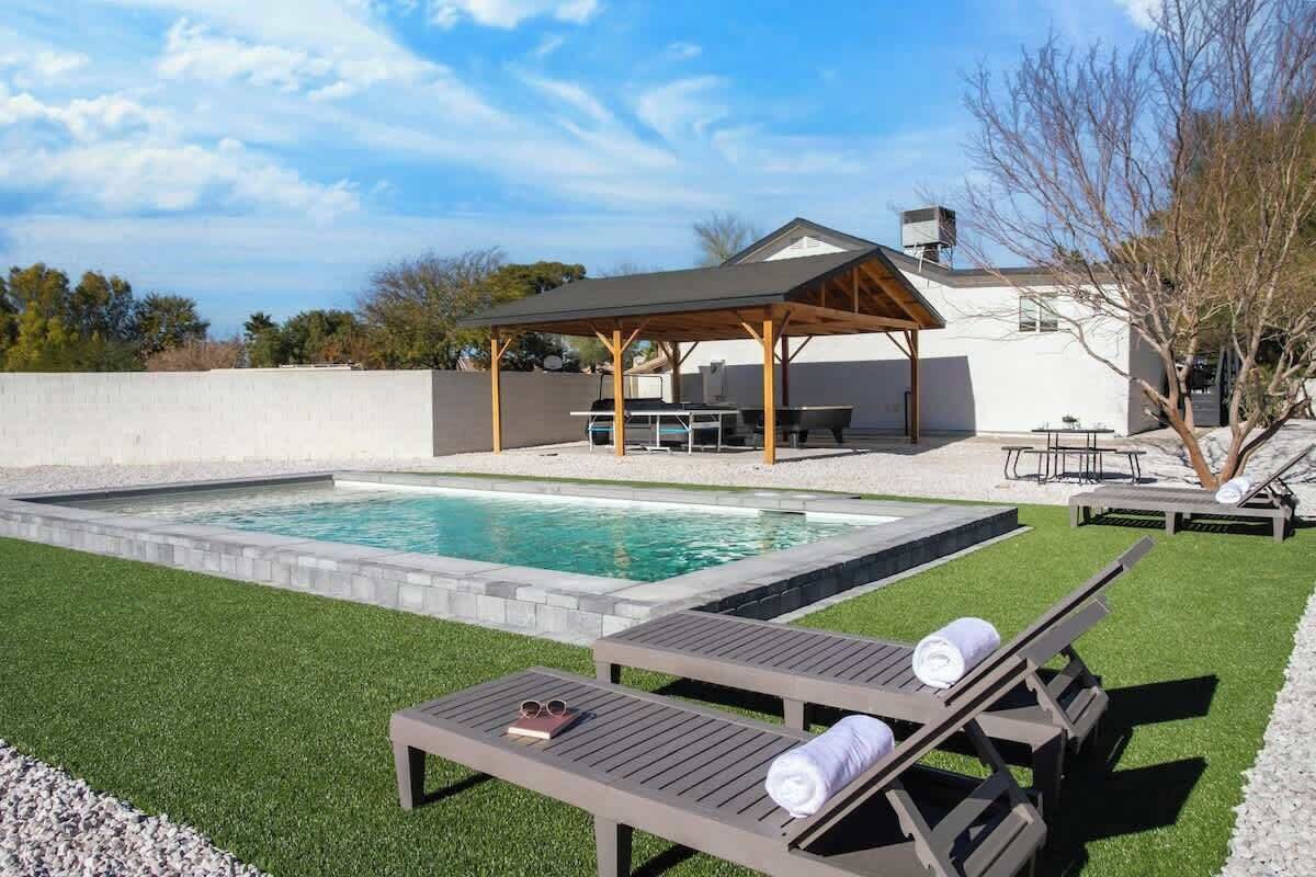 A backyard with a swimming pool, two lounge chairs, a covered patio, and a barbecue area.