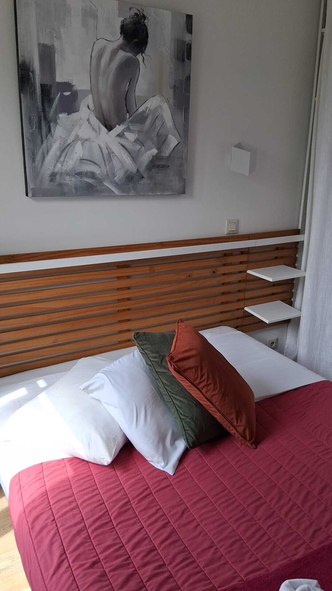 Une chambre à coucher avec un lit, des oreillers, une tête de lit en bois, un tableau au mur et une fenêtre avec des rideaux.