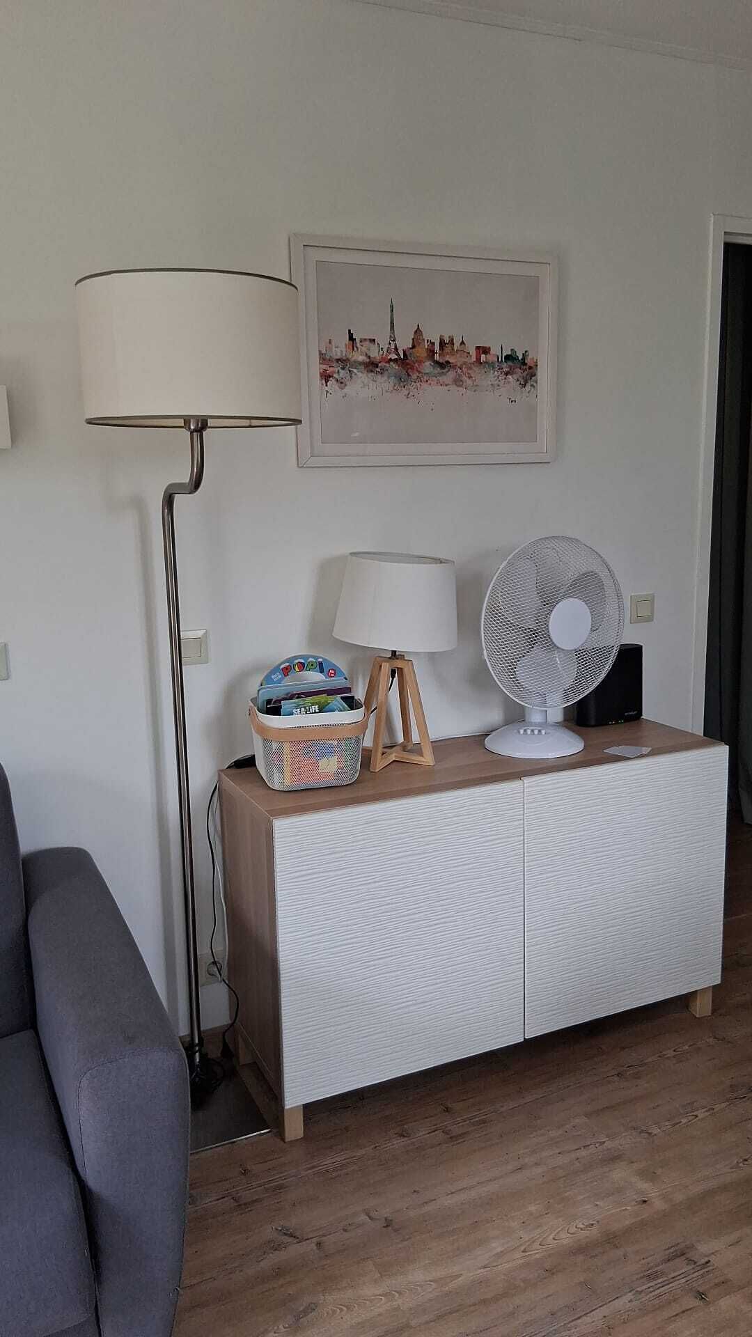 A room with a white cabinet, a floor lamp, a desk lamp, a fan, and a framed cityscape picture on the wall.