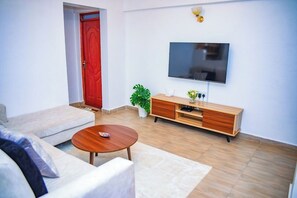 A living room with a sofa, coffee table, TV, wooden cabinet, and a red door. - Lux Suites Father's Hub Apartments (Nairobi)