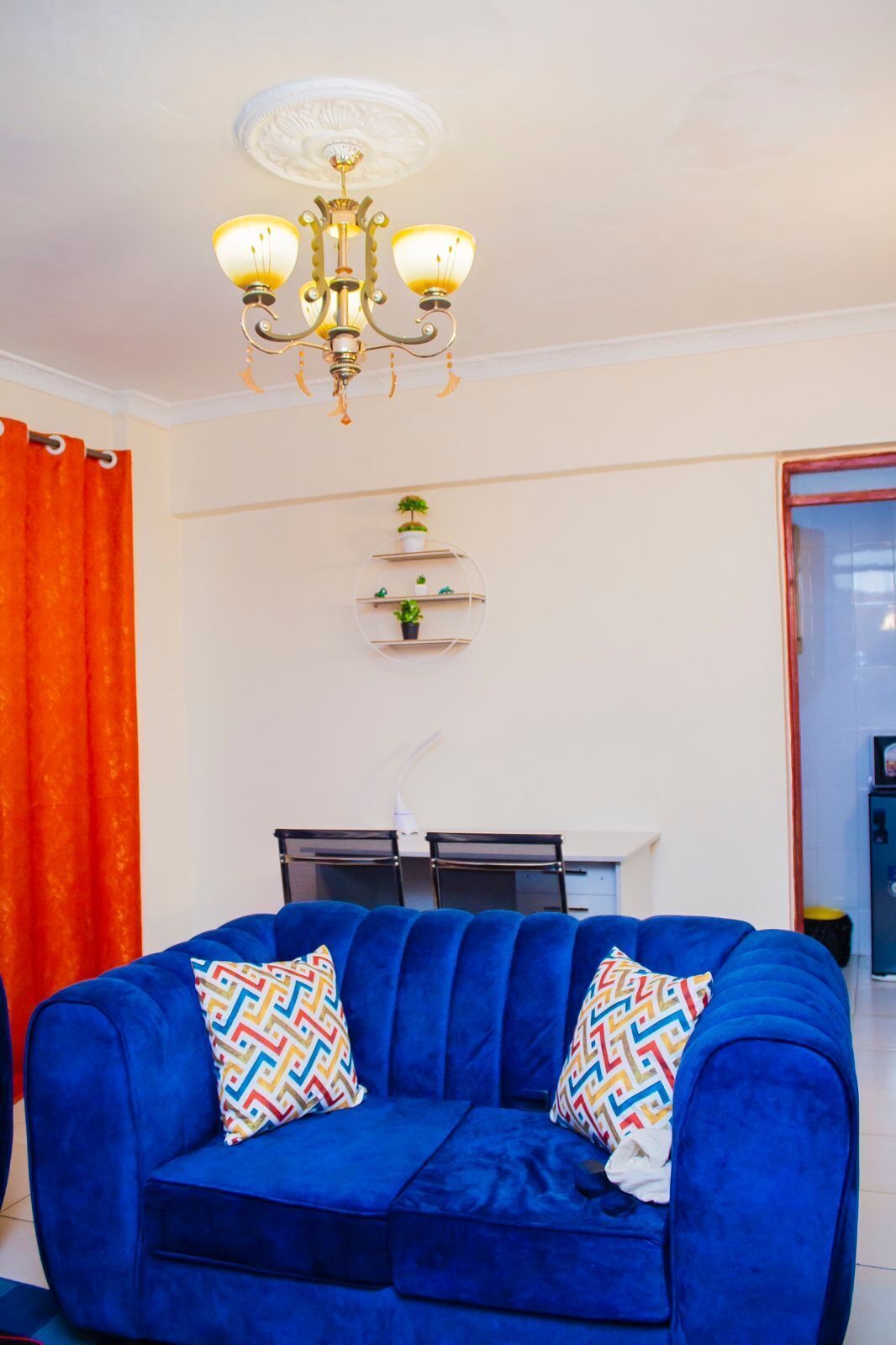 A living room with a blue sofa, a chandelier, a small plant shelf, and a dining area in the background.