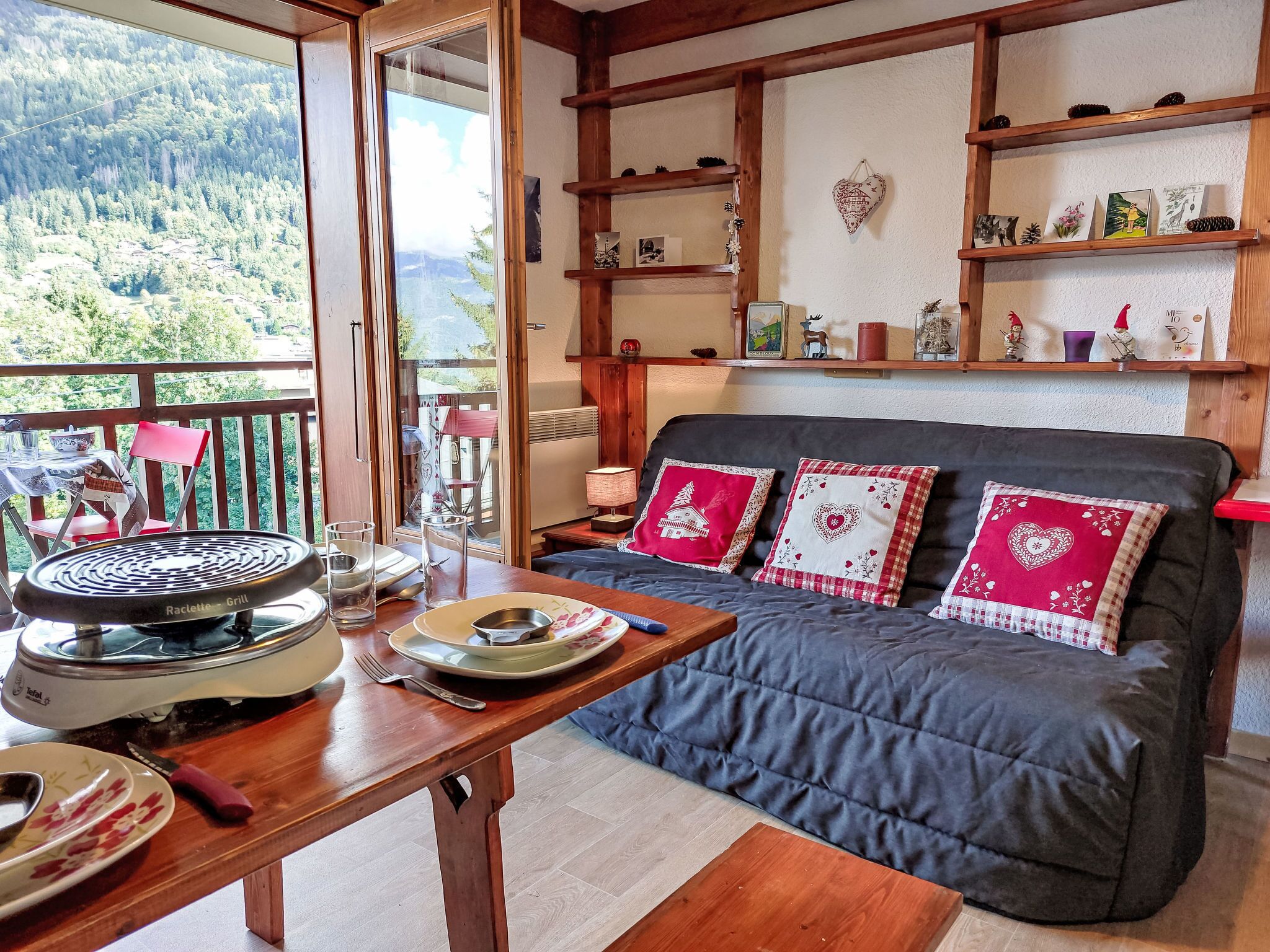 A cozy living room with a dark blue sofa, wooden shelves with decorative items, a dining table with plates and glasses, and a view of a forested landscape through a large window.