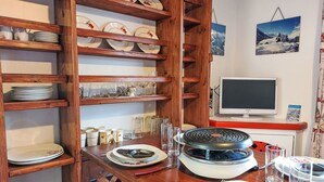 Uma área de refeições com prateleiras de madeira que exibem pratos e copos, uma churrasqueira sobre a mesa e uma televisão na parede.