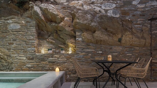 A stone wall with a built-in seating area, a pool, and a table with chairs. - Porto Sereno Andros Suites (Andros)