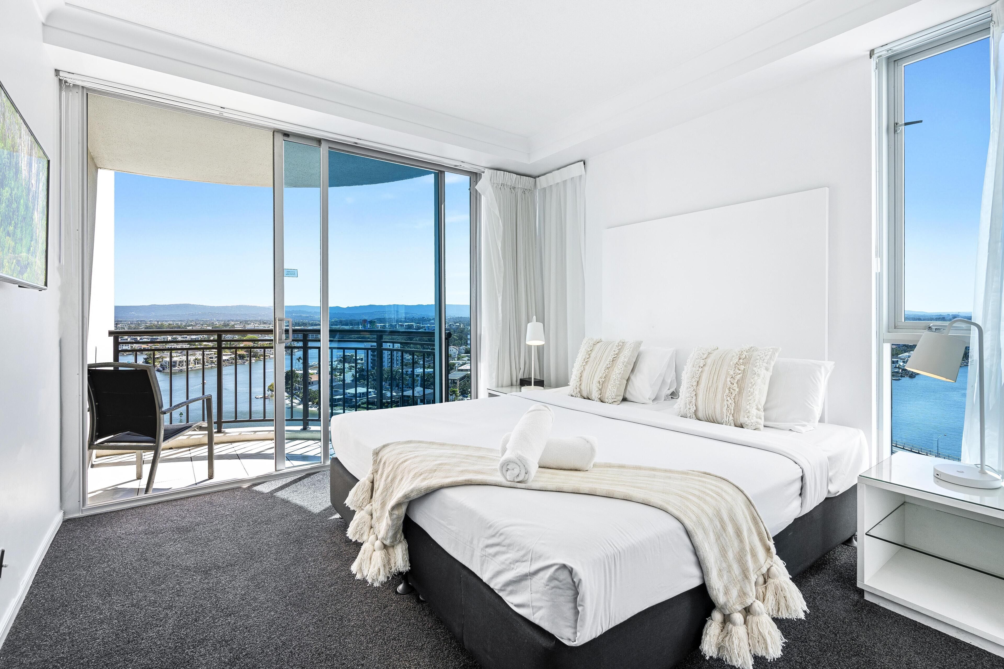 A modern hotel room with a large bed, a balcony with a view of the city, and a cityscape visible through the window.