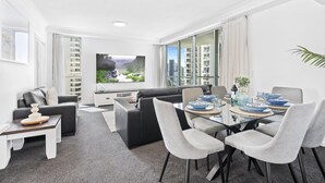 A modern living room with a glass dining table, grey and black sofas, and a large TV.