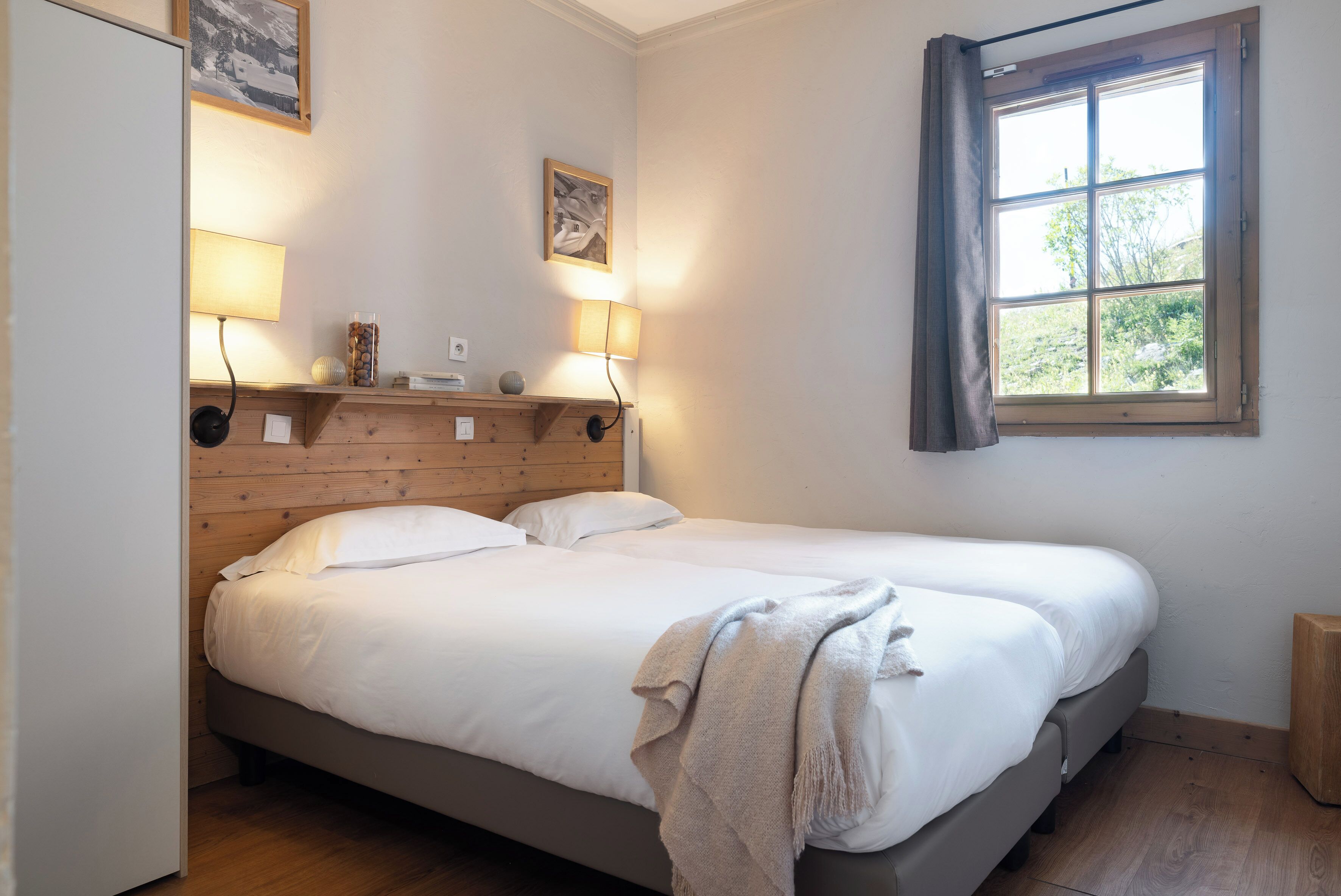A bedroom with two single beds, wooden headboard, bedside lamps, a window with curtains, and a small shelf with decorative items.