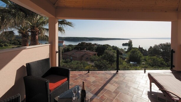 Ein Balkon mit Korbmöbeln, einem Tisch mit Flasche und Gläsern sowie Blick auf einen See und Gebäude.