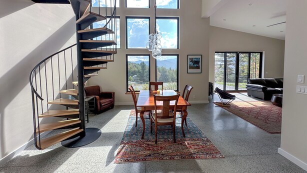 Un vaste espace de vie comprenant une table à manger en bois, des chaises et un tapis à motifs. Un escalier en colimaçon mène à l’étage supérieur. De grandes fenêtres offrent une vue sur l’extérieur.