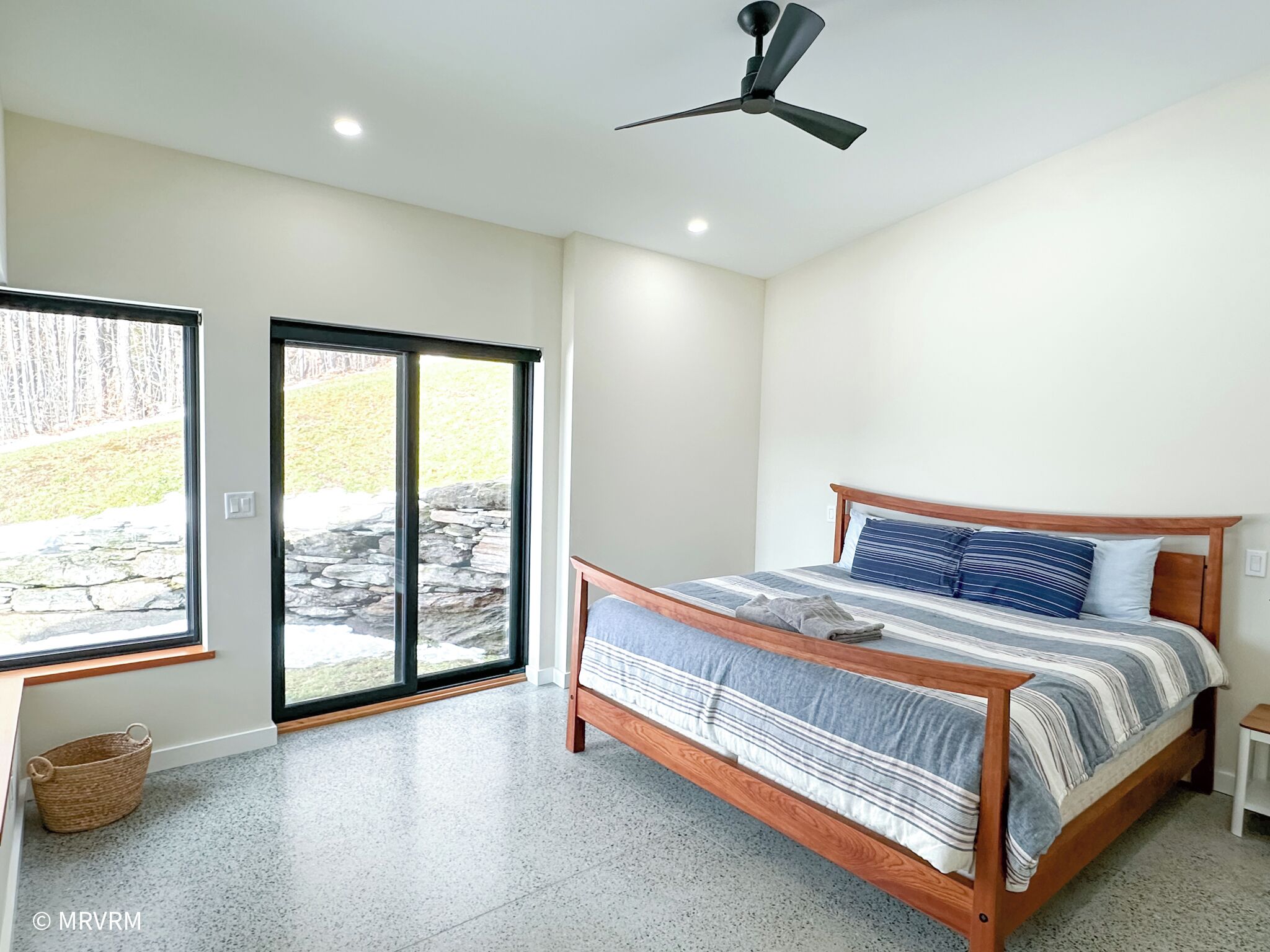 Un dormitorio con cama, ventilador de techo, ventana con vista a una pared de piedra y follaje, y una cesta en el suelo.