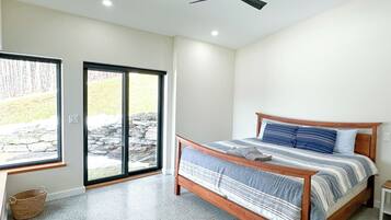 Un dormitorio con cama, ventilador de techo, ventana con vista a una pared de piedra y follaje, y una cesta en el suelo.