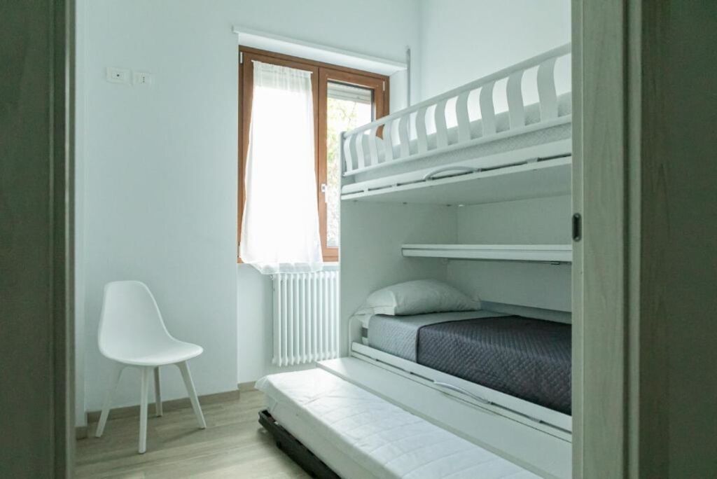 A small, single-room apartment with a bunk bed, a white chair, and a window with a curtain.